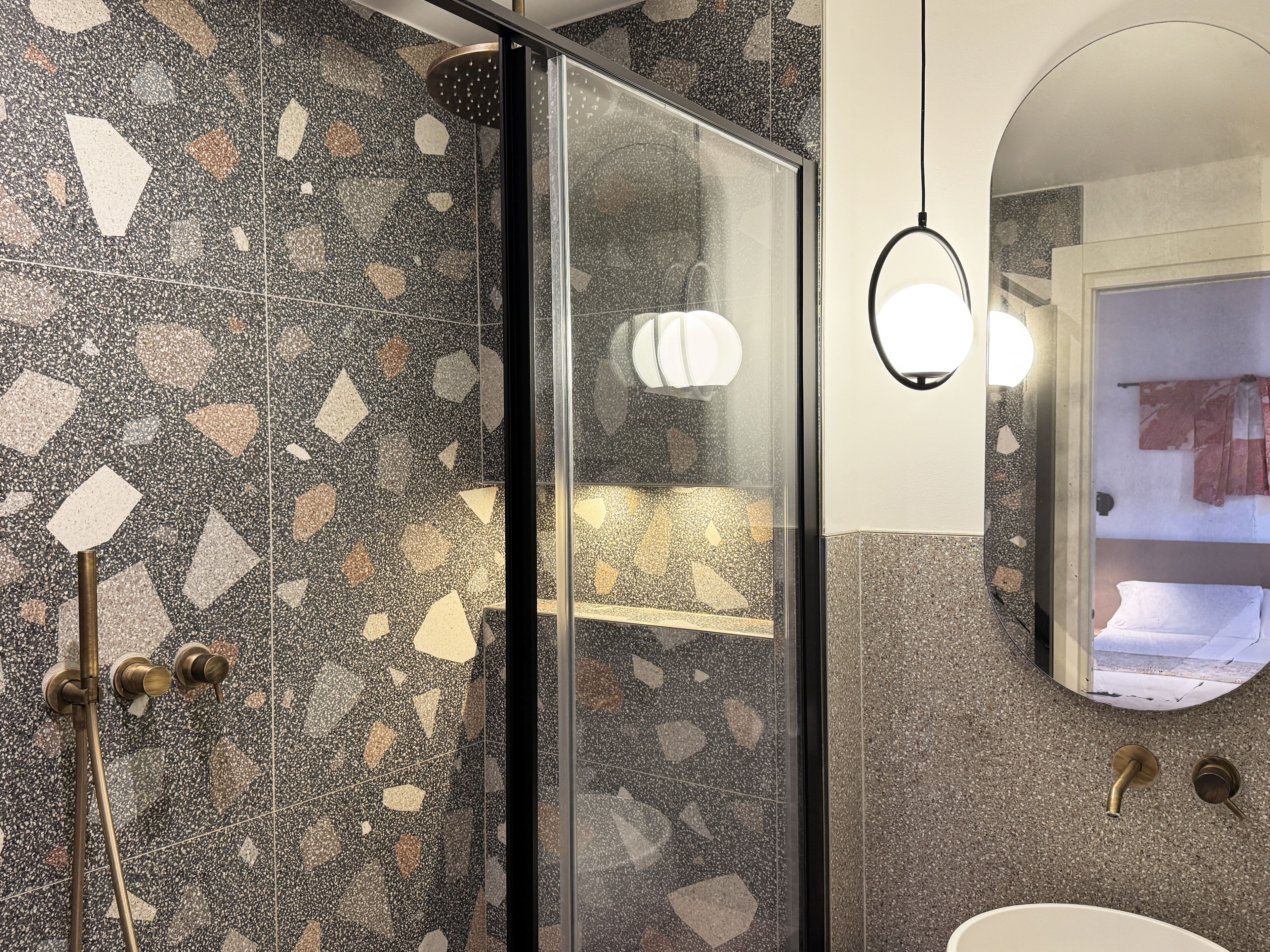 A modern bathroom with a mosaic tile floor, a glass shower enclosure, a round mirror, and a sink with a gold faucet.