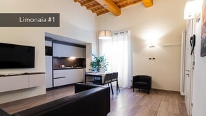 A modern living room with a black sofa, a flat-screen TV, a dining area with a table and chairs, and a kitchen in the background. - Limonaia 1 | Apartment Villa Carobbi (Firenze)