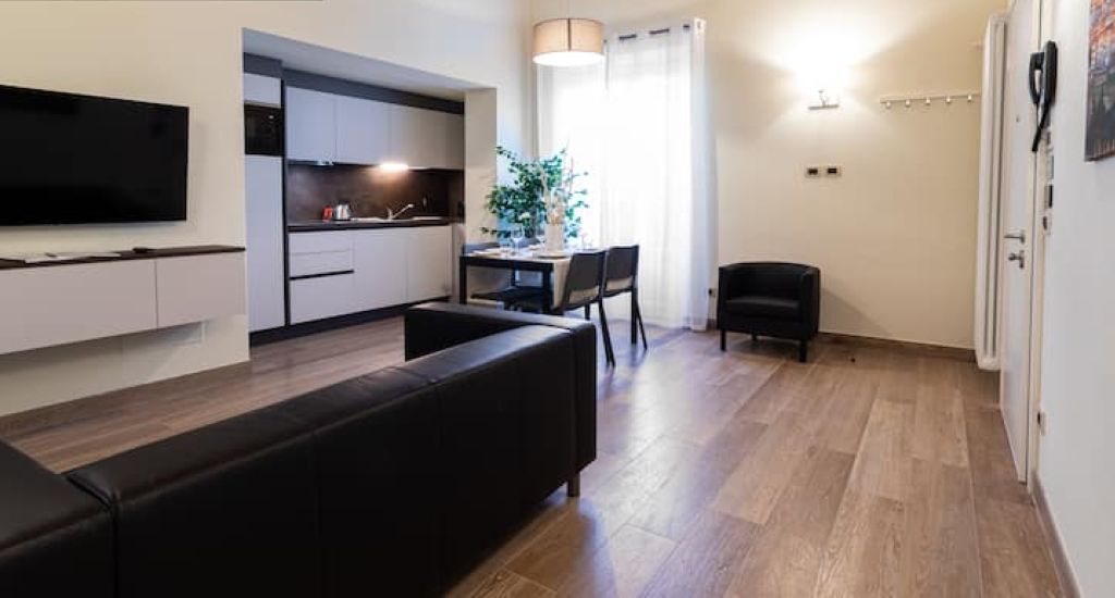 Ein modernes Wohnzimmer mit einer schwarzen Couch, einem Flachbildfernseher, einem Essbereich mit Tisch und Stühlen sowie einer Küche im Hintergrund.