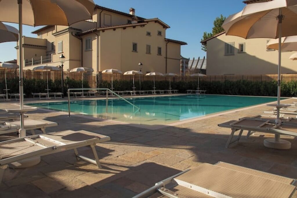 A pool area with lounge chairs and umbrellas, surrounded by a wooden fence and buildings.