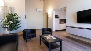 A modern living room with a wooden ceiling, a black sofa, two black armchairs, a coffee table with a magazine, a potted plant, and a TV mounted on the wall. - Ancient Well 3 | Villa Carobbi Apt (Firenze)