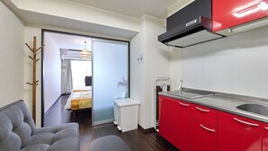A compact kitchen with red cabinets, a grey sofa, and a small white trash bin. - XS16 XROSS STAY Namba8 (大阪市中央区島之内)