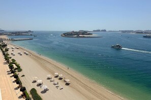 Una playa con filas de tumbonas y sombrillas, un barco en el agua, y una vista distante de un horizonte urbano.