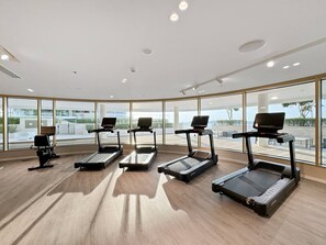 Une salle de sport moderne avec cinq tapis de course alignés en rangée, de grandes fenêtres donnant sur l’extérieur et un plancher en bois.