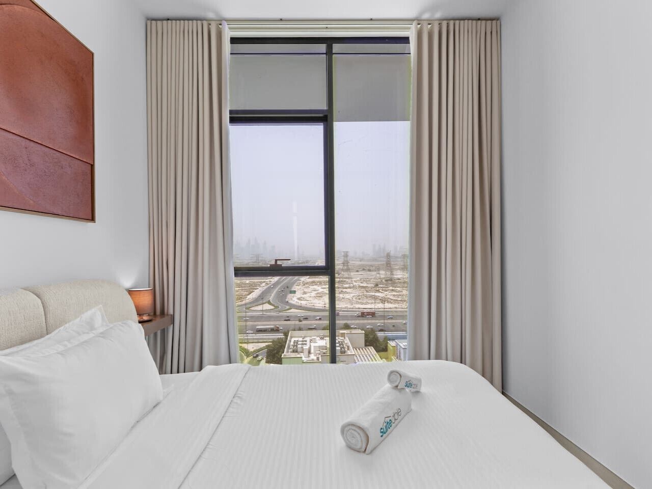 Une chambre d’hôtel avec un grand lit, une table de chevet, une lampe et une vue sur la ville, visible à travers une fenêtre avec des rideaux.