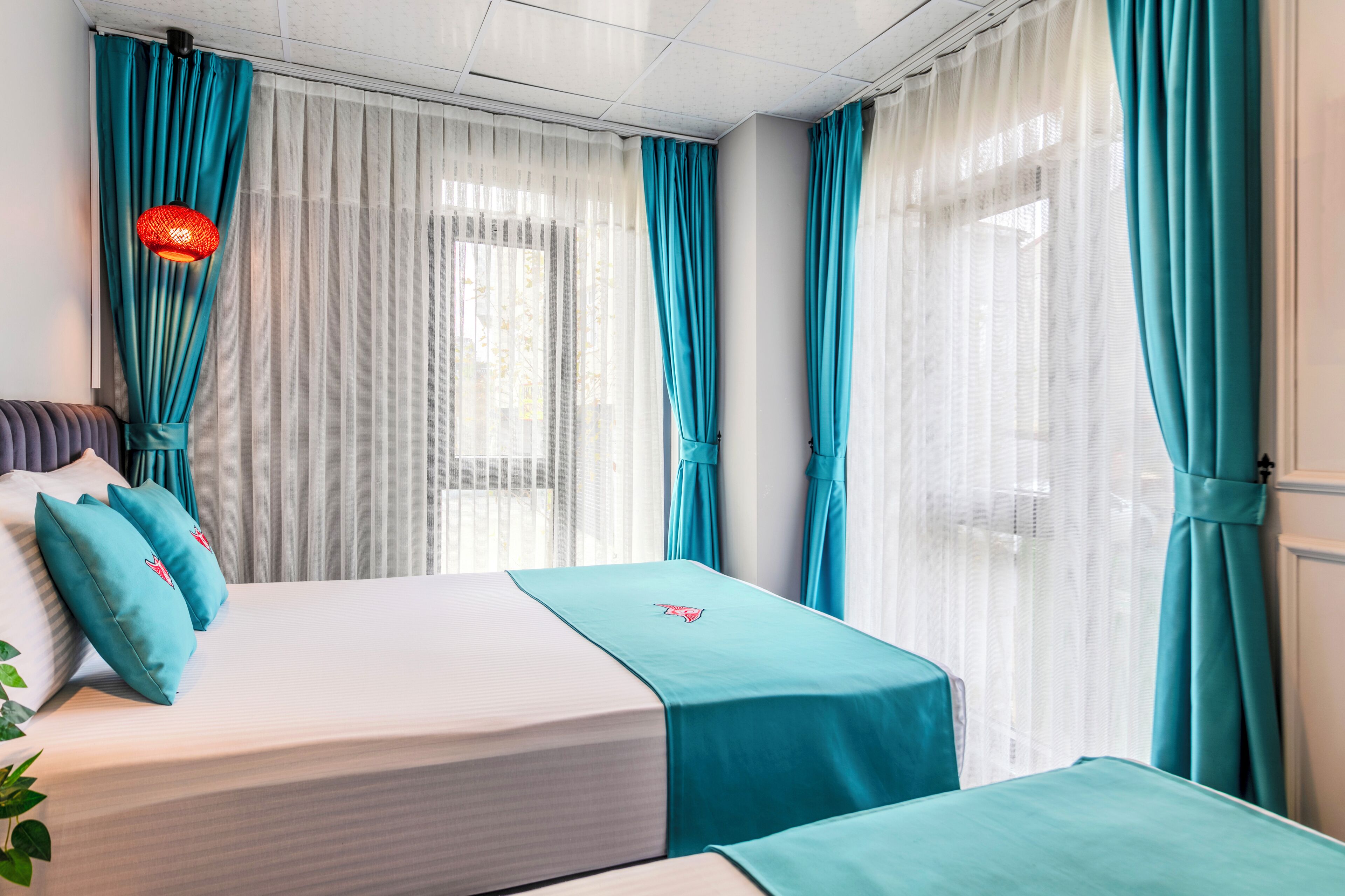 Une chambre d’hôtel avec deux lits, des rideaux turquoise, un luminaire suspendu rouge et une fenêtre ornée de rideaux blancs et légers.