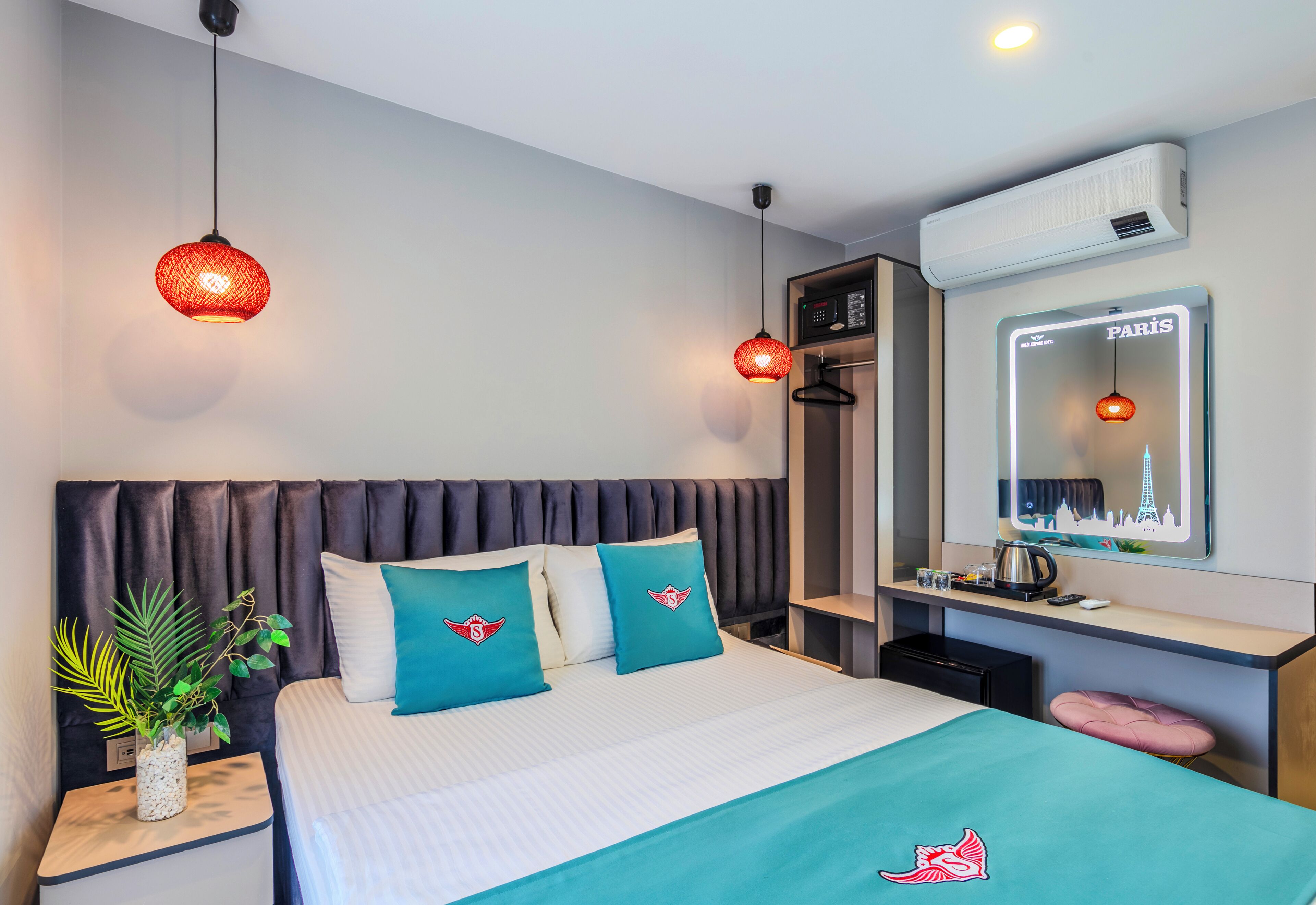 A hotel room with a bed, two red hanging lamps, a potted plant, a minibar, and a mirror with a cityscape reflection.