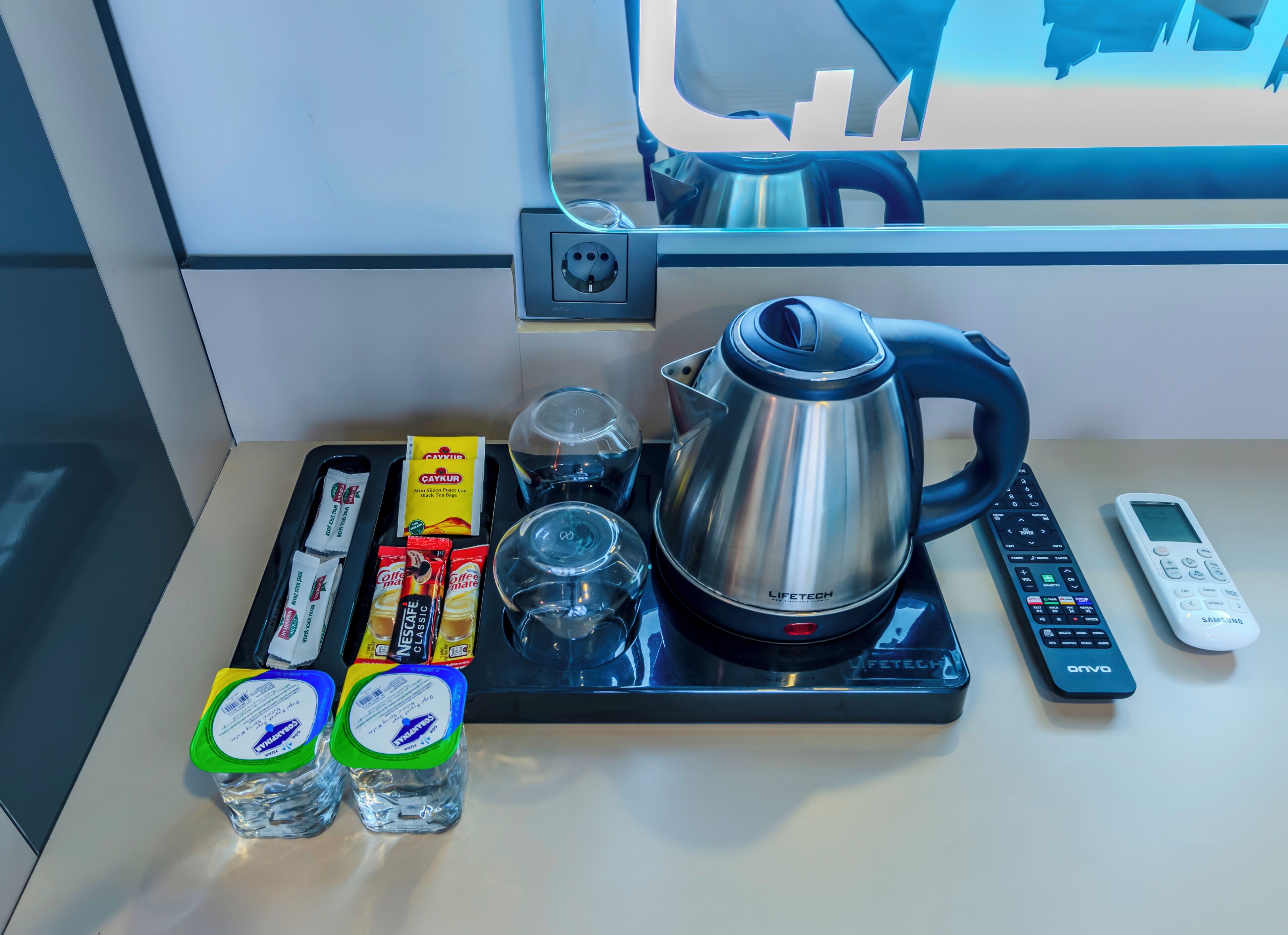 Un plan de travail de cuisine avec une bouilloire, une machine à café, une télécommande, un téléphone intelligent et deux bouteilles d’eau.
