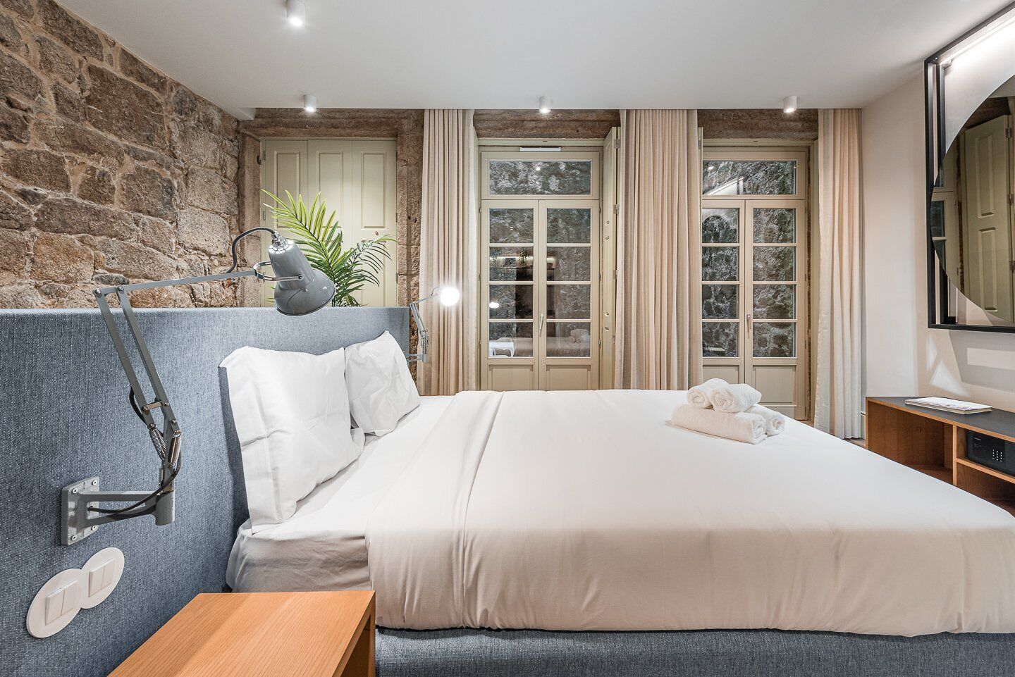 Une chambre moderne avec un mur en pierre, un grand lit, une table de chevet, une lampe et une fenêtre avec des rideaux.
