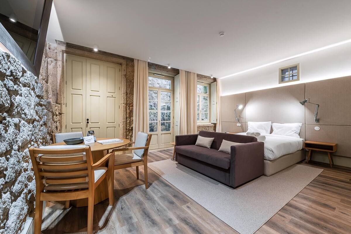A modern room with a wooden dining table, a sofa, a bed, and a stone wall.