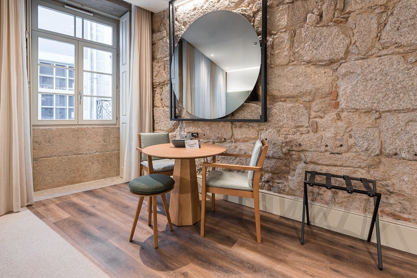 Une pièce avec un mur en pierre, une table ronde en bois accompagnée de deux chaises, un miroir et une fenêtre avec des rideaux.