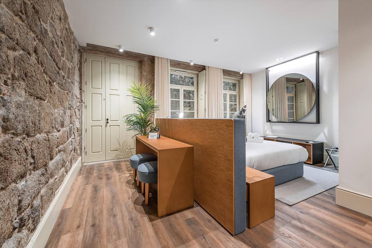A bedroom with a bed, a desk, and a chair, a large mirror, and a stone wall.