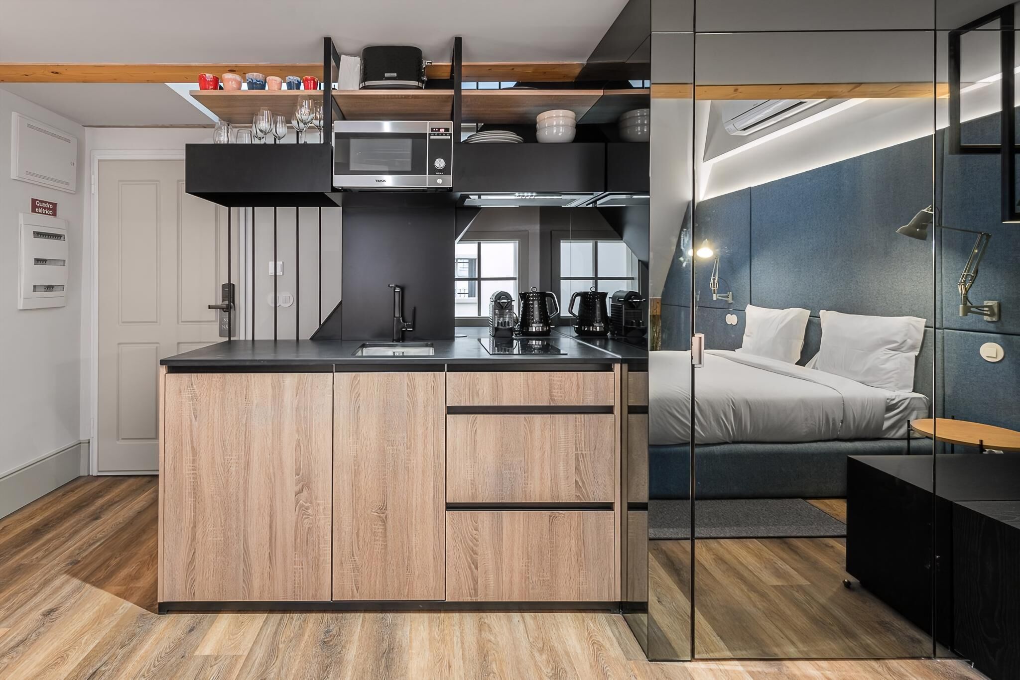 A modern kitchen with a wooden island, built-in appliances, and a glass partition separating the kitchen from a bedroom with a bed and a nightstand.