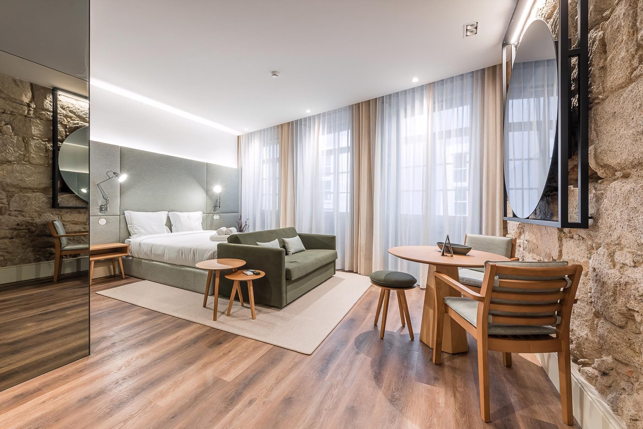 Ett modernt hotellrum med en vägg som har en dekorativ stenbeklädnad, en stor säng, en grön soffa, en matplats med bord och stolar samt en stor spegel.