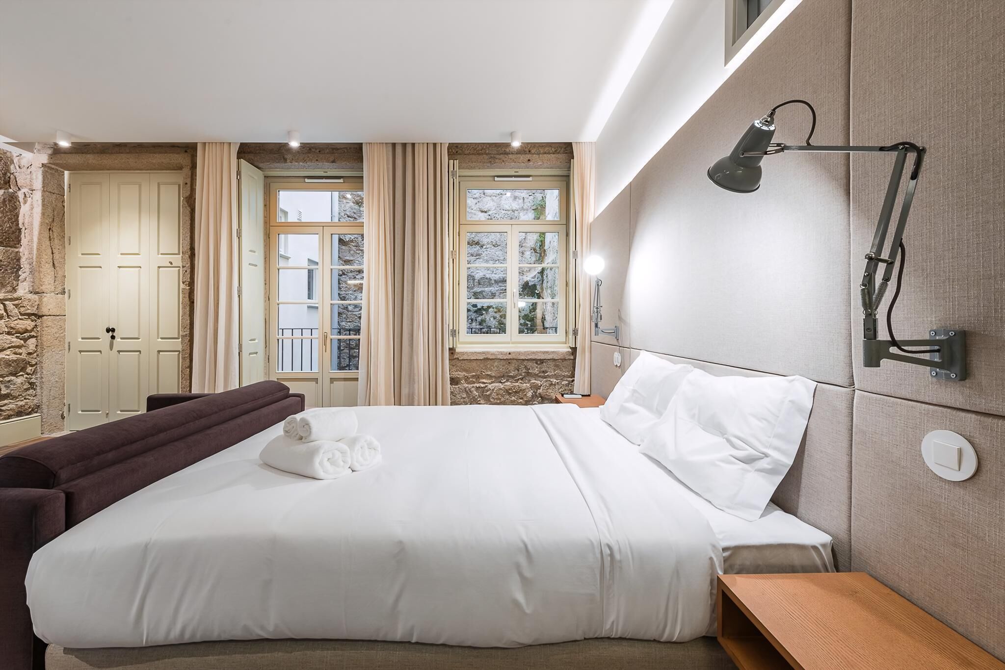 Una cama bien tendida con ropa blanca, una mesita de noche, una lámpara de pared y vistas a un muro de piedra y una ventana con cortinas.