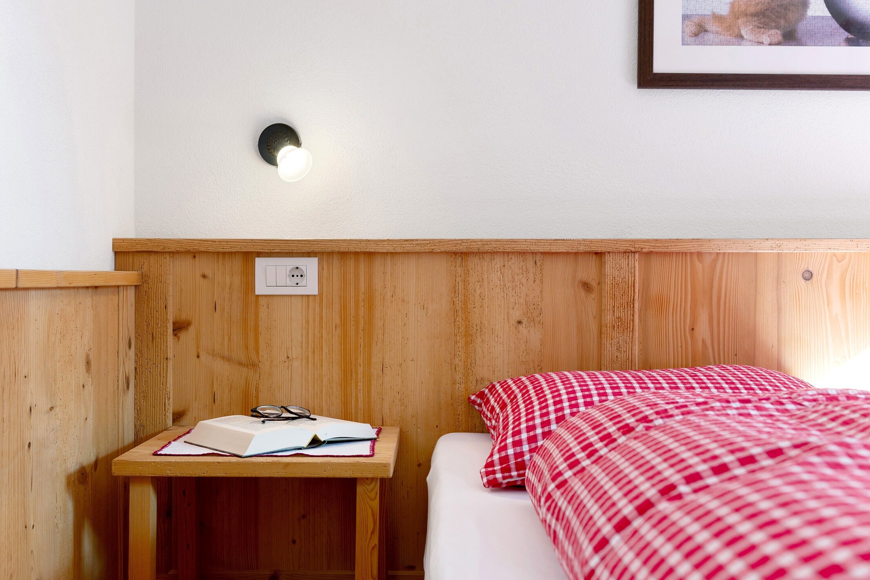 A wooden headboard, a bedside table with a book and glasses, a red and white checkered bedspread, and a wall-mounted light fixture.