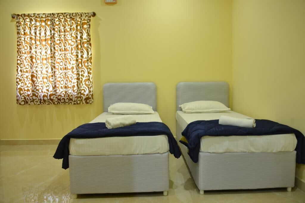 Dos camas individuales con ropa de cama blanca y azul, una cortina estampada y una pared amarilla.