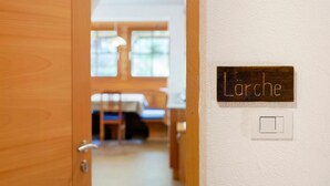 Ein Hotelzimmer-Eingang mit Holztür, einer Namensschild mit der Aufschrift 'Larché', einem Lichtschalter und einem Blick auf ein Bett und ein Fenster im Hintergrund.