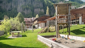 Ein Spielplatz mit Rutsche und Klettergerüsten, umgeben von Gebäuden im Holzbau und einem bewaldeten Berg im Hintergrund.