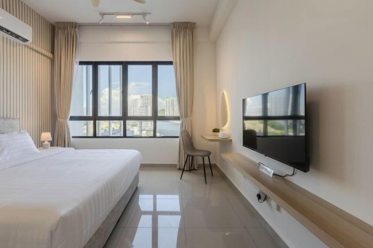 A hotel room with a bed, a TV mounted on the wall, a desk with a chair, and a window with a view of buildings.