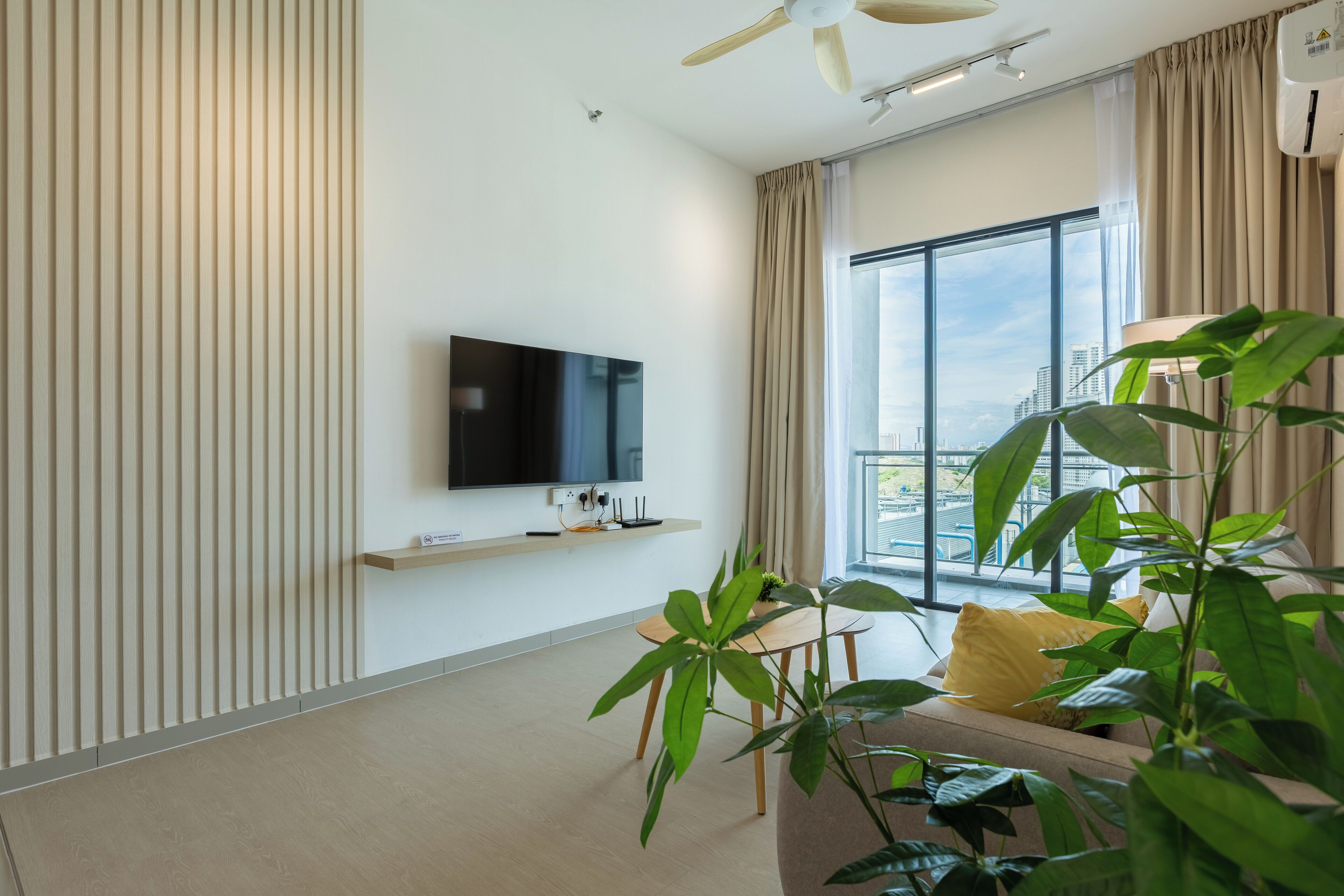 A modern living room with a flat-screen TV mounted on the wall, a dining table with chairs, and a large window offering a city view.
