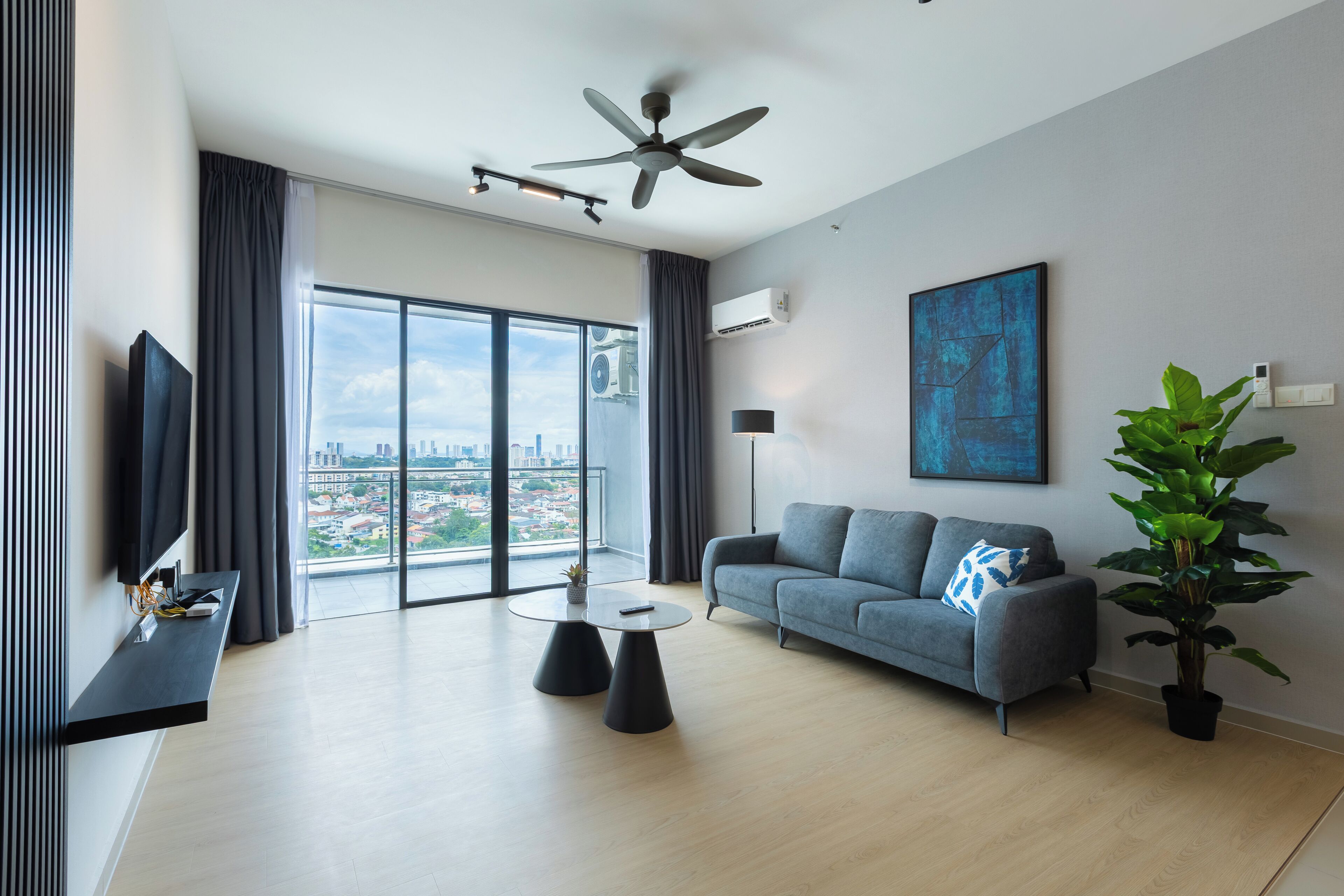 Un soggiorno moderno con un divano grigio, un tavolino rotondo, una ventola a soffitto e un'ampia finestra con vista sulla città.