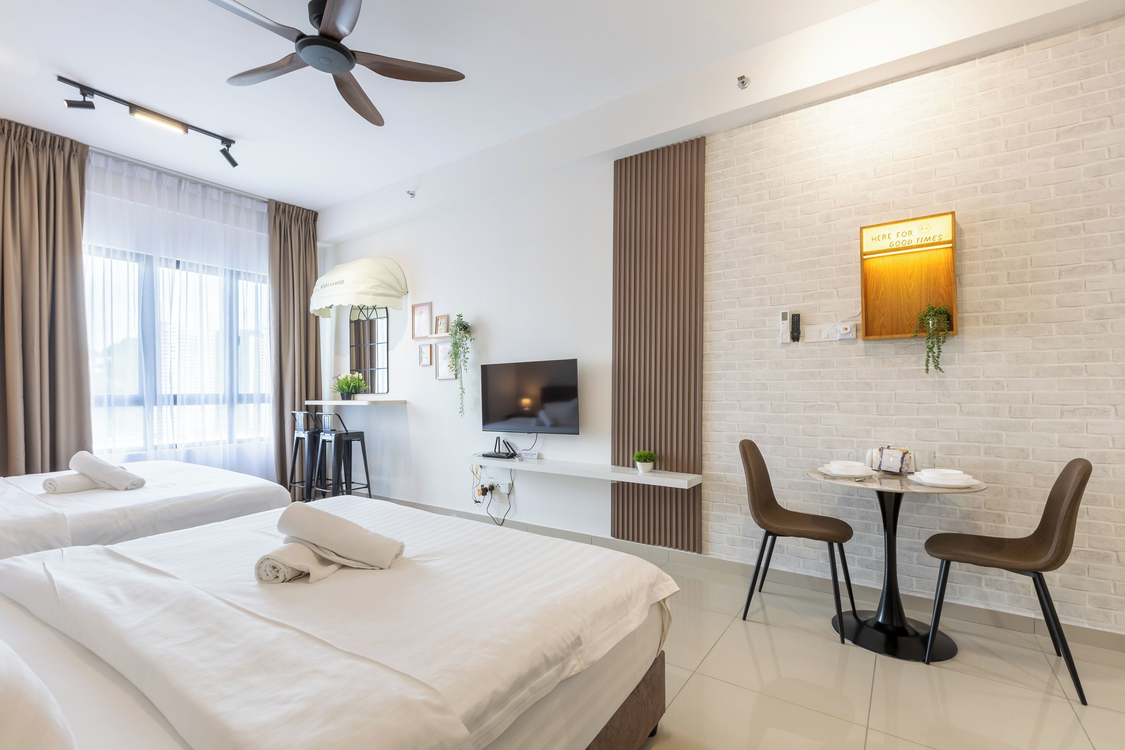 Habitación de hotel con dos camas, televisión, mesa de comedor con sillas, ventilador de techo y pared de ladrillo con un cartel.