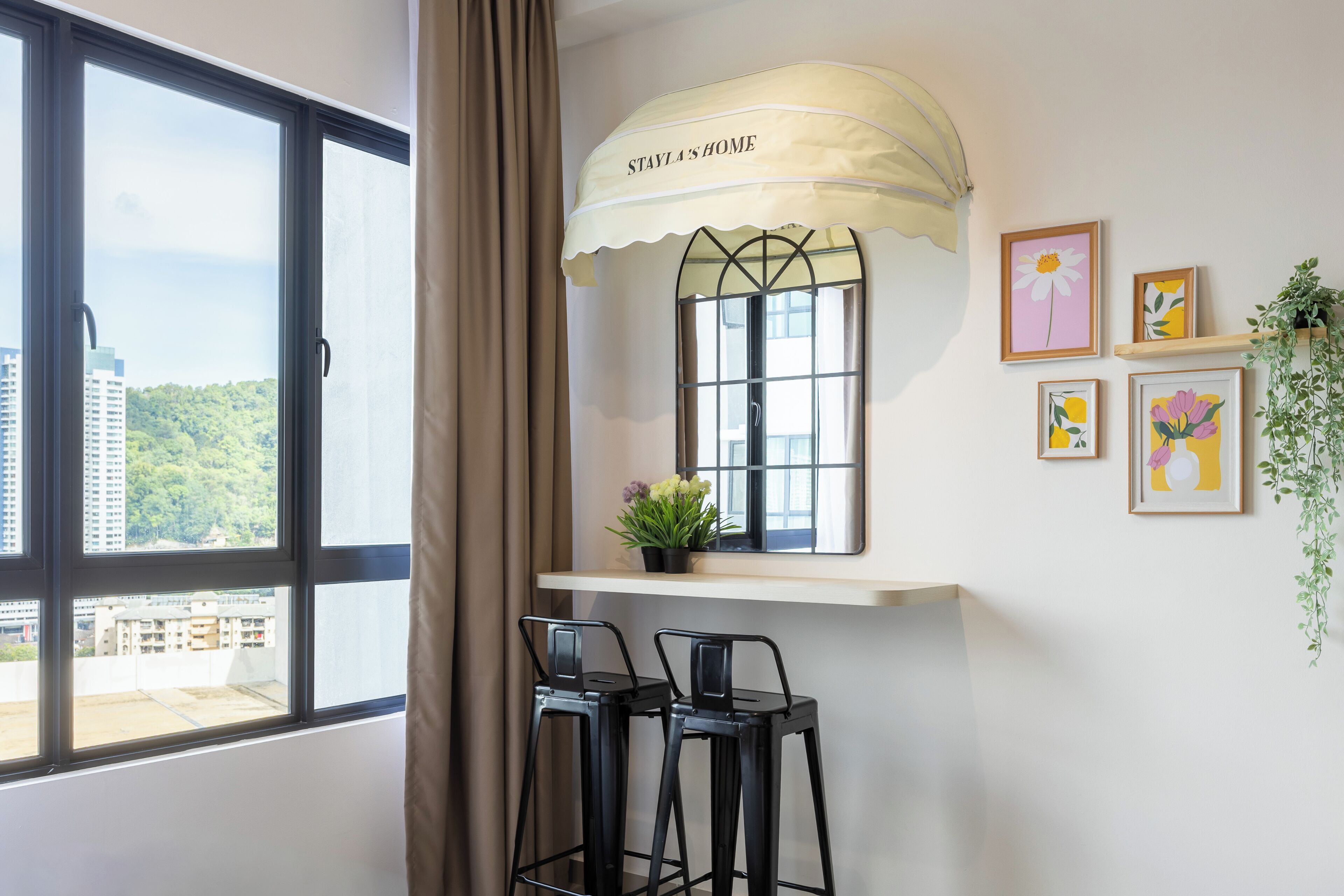 A room with a large window, a small table with two chairs, a potted plant, and framed artwork on the wall.