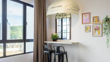 A room with a large window, a small table with two chairs, a potted plant, and framed artwork on the wall.