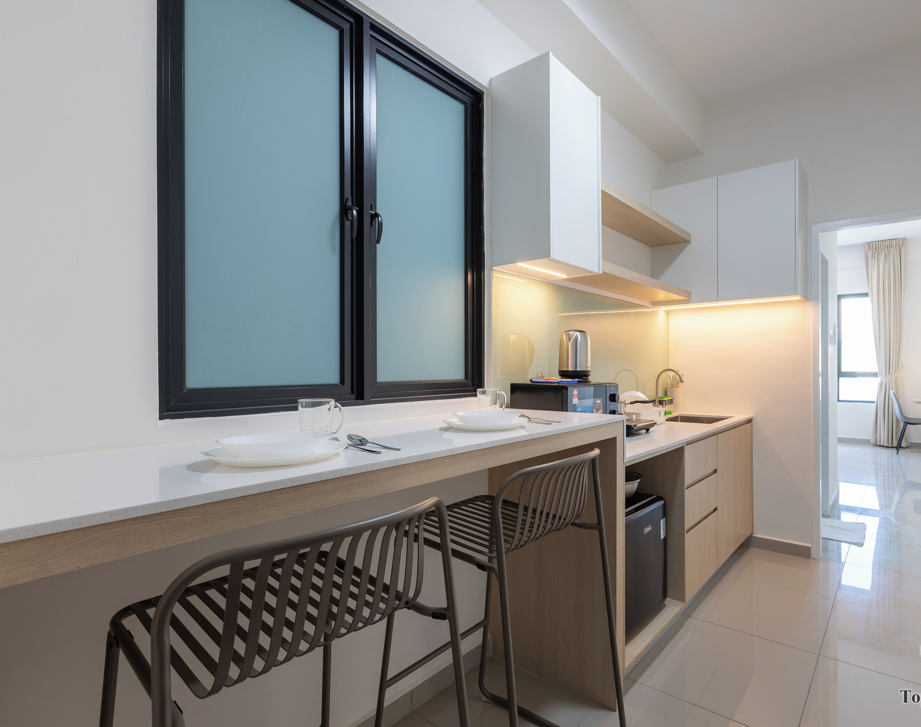 A modern kitchen with a large window, white cabinets, and a built-in oven. There are two bar stools and a small sink.