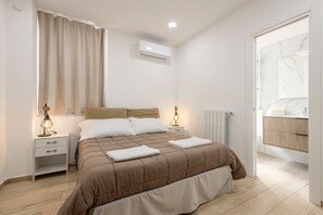 Un dormitorio moderno con cama, mesitas de noche, aire acondicionado, una ventana con cortinas y un baño visible a través de una puerta abierta.