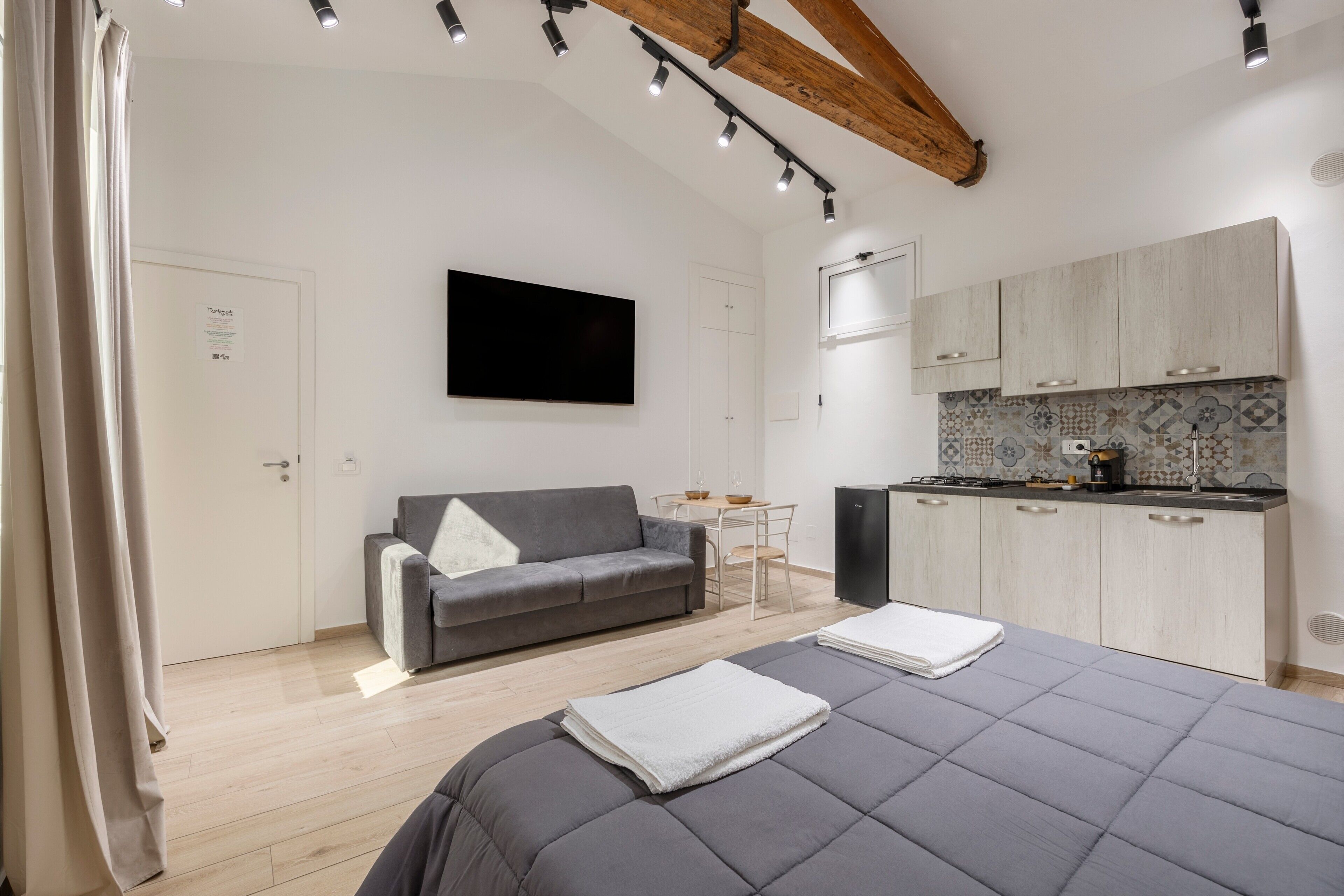 Une chambre moderne comprenant un lit, un canapé gris, une petite table à manger, un coin cuisine avec des placards et un téléviseur fixé au mur.