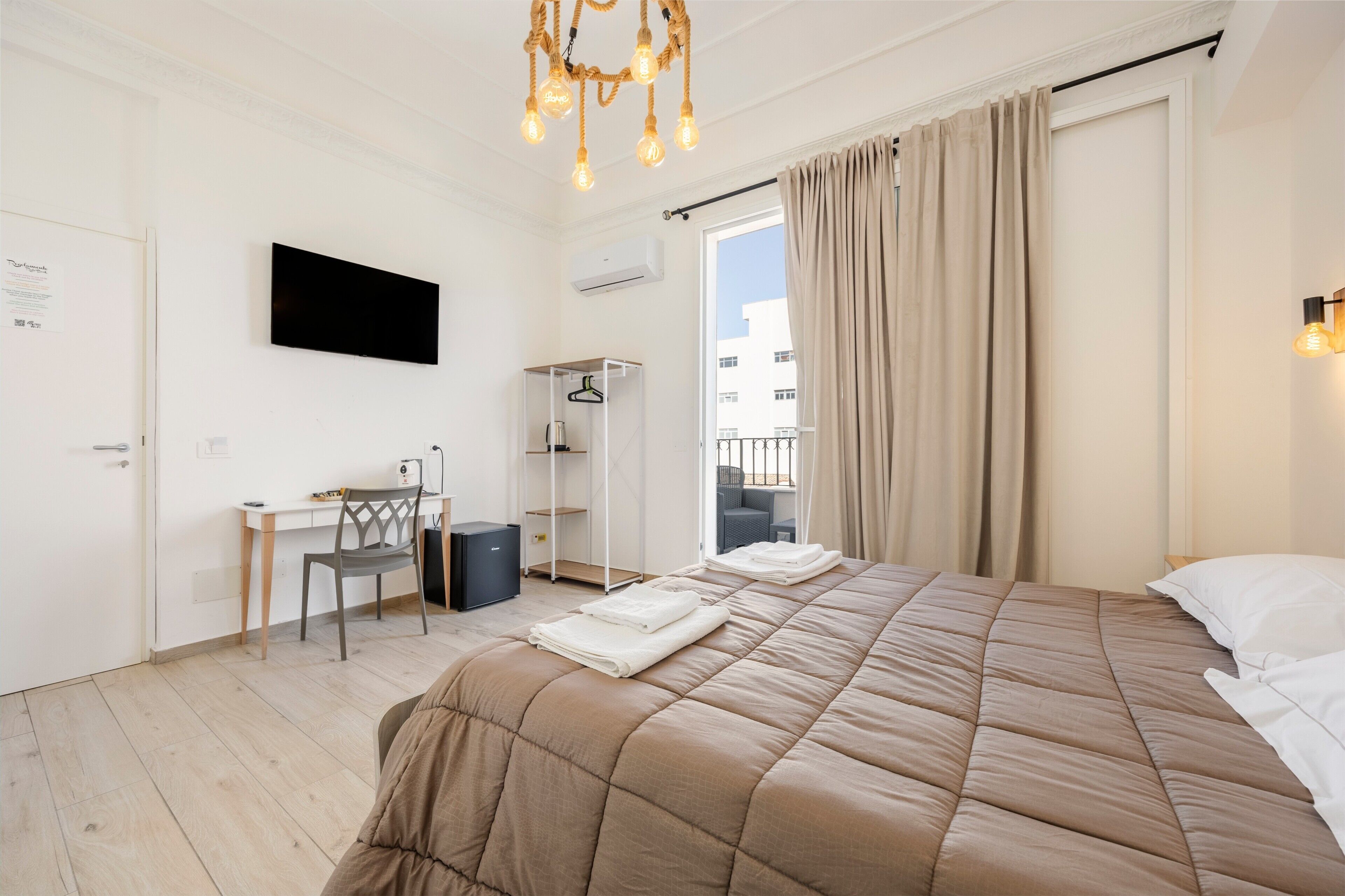 A bedroom with a bed, a desk, a chair, a TV, and a balcony with curtains.