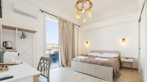 A modern hotel room with a bed, a desk, a chair, a balcony with a view, and a chandelier. - Mangano Suite Apartment (Palermo)
