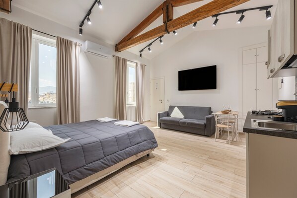 Un dormitorio moderno con cama, un sofá gris, una pequeña mesa de comedor y una zona de cocina.
