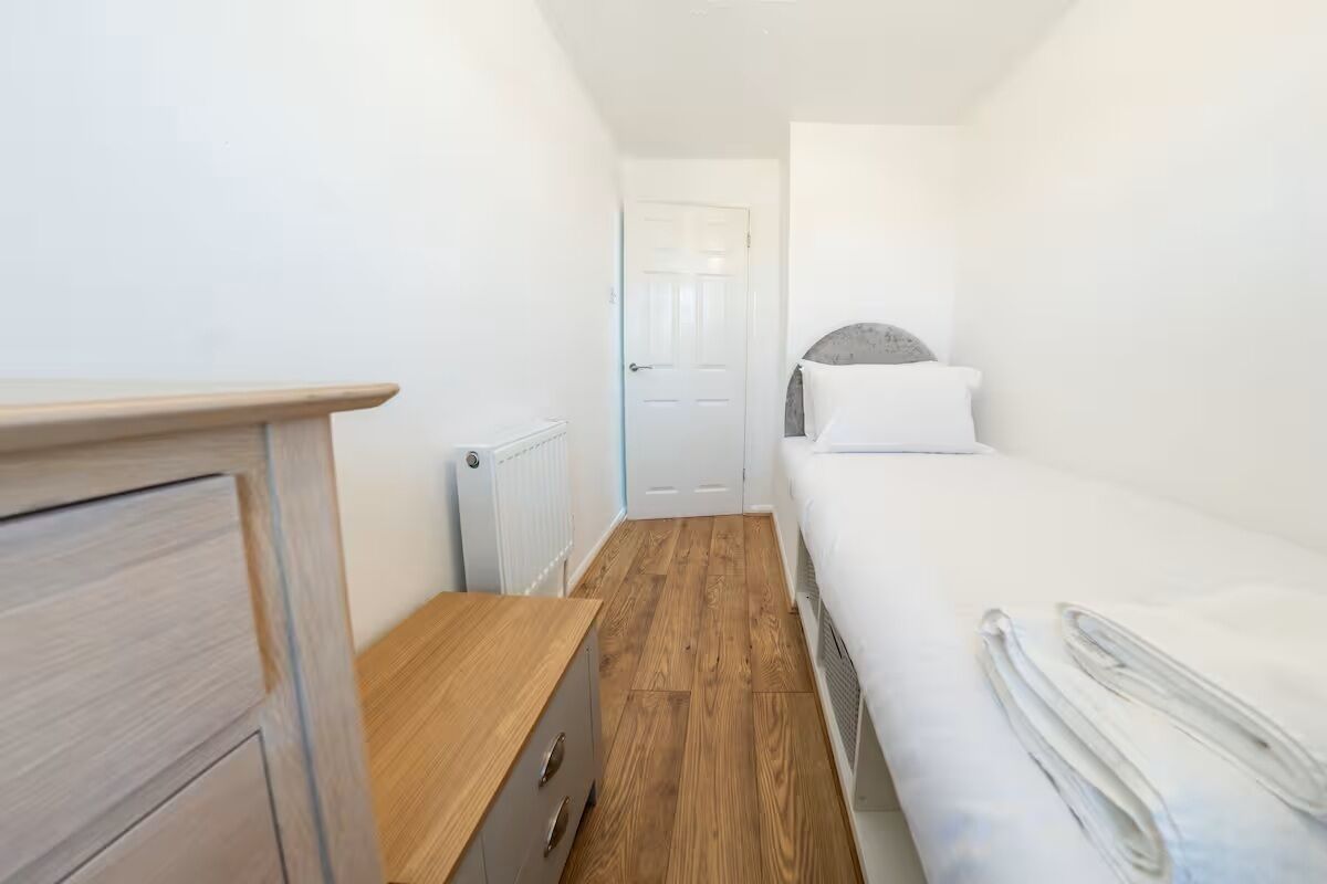 Una habitación pequeña y estrecha con un aparador de madera, una cama con ropa de cama blanca y un radiador en el suelo.