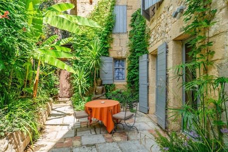 A stone courtyard with a round table and chairs, surrounded by lush greenery and a Buddha statue.