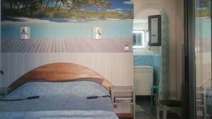 A bedroom with a bed, a nightstand, a small table, a mirror, and a view of a beach scene. - Large T2 in villa with sea view and spa not overlooked (La Ciotat)
