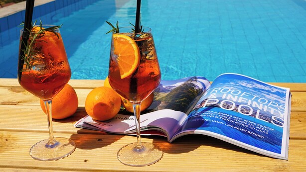 Una mesa de madera con dos vasos de té helado, una revista y naranjas junto a una piscina.