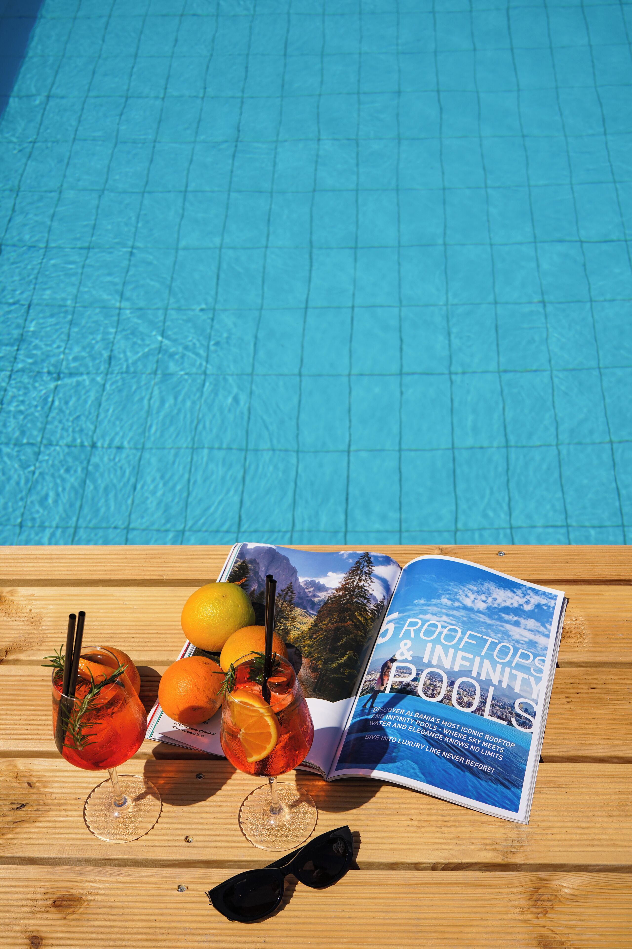 A magazine titled "ROOFTOPS & INFINITY POOLS" on a wooden table by a pool.