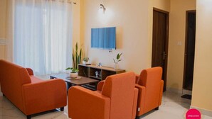 A living room with orange sofas, a coffee table, a TV mounted on the wall, and a door leading to another room.