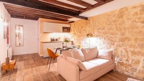 Una sala de estar acogedora con pared de piedra, techo de madera, un sofá color beige y una cocina al fondo.