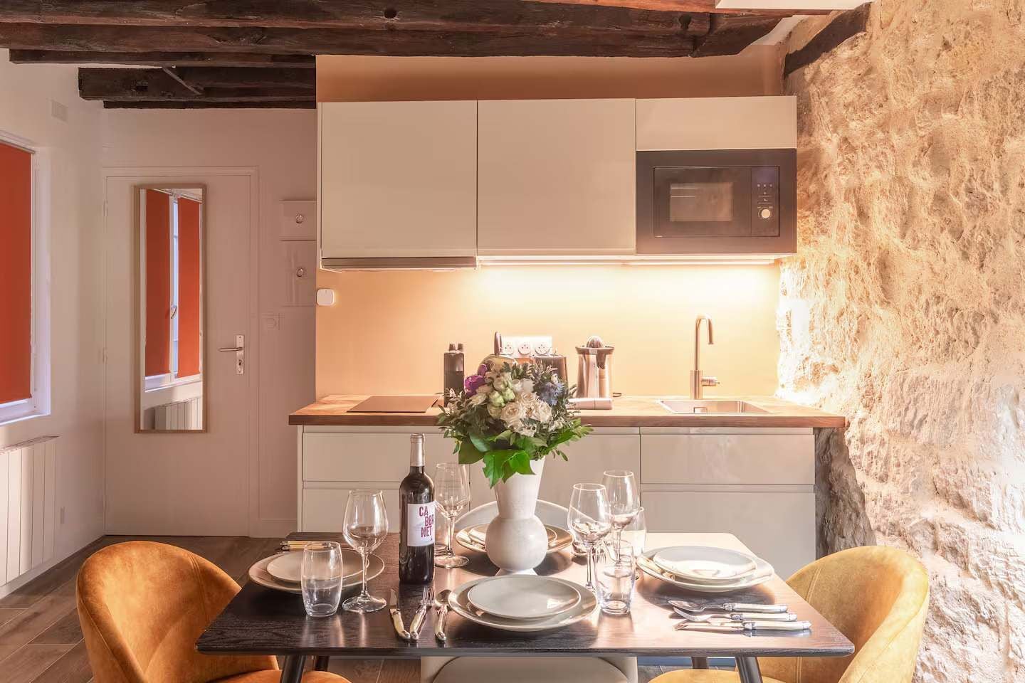 A dining area with a table set for four, a vase with flowers, and a bottle of wine. A kitchen is visible in the background with a microwave and a sink.