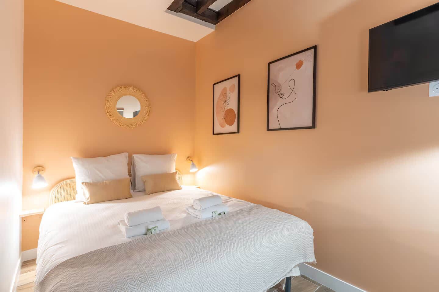 A hotel room with a bed, a round mirror, two framed artworks, and a flat-screen TV.