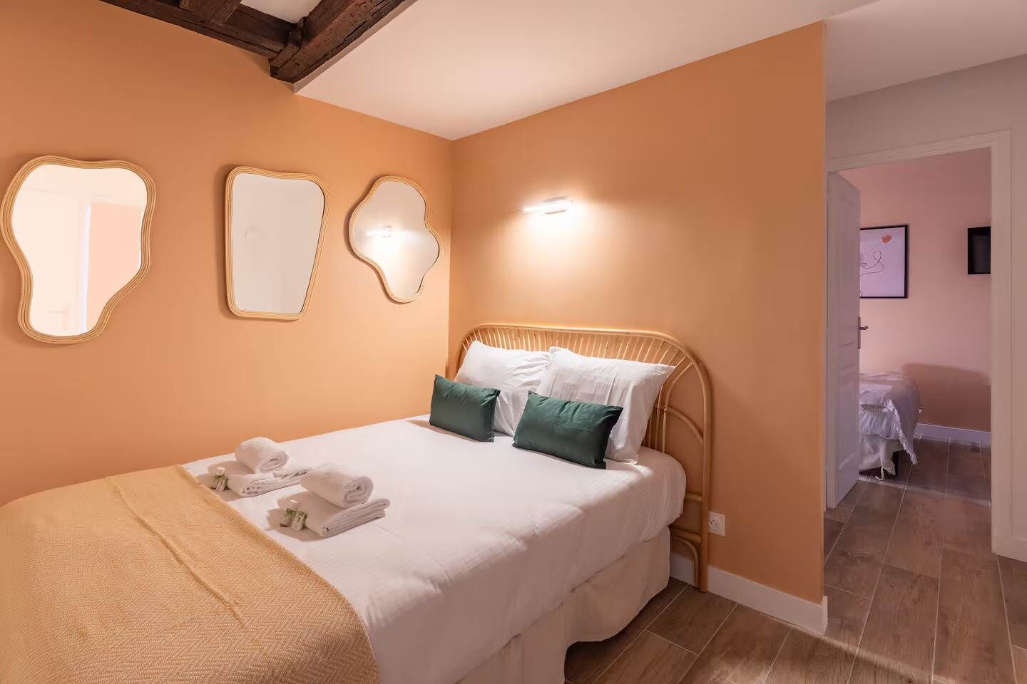 Un lit bien fait, avec du linge de lit blanc et deux coussins verts, une tête de lit en bois et un miroir fixé au mur.