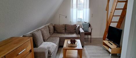 Un salon chaleureux avec un canapé d’angle, une table basse en bois, un téléviseur posé sur un meuble, un bureau avec une chaise et une échelle en bois.