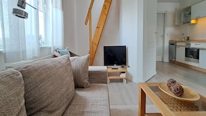Una sala moderna con un sofá, un televisor sobre un mueble, una escalera de madera y una cocina con armarios y electrodomésticos.