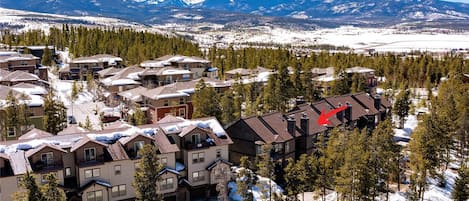 Un barrio residencial con varias casas, algunas con nieve en los tejados, rodeado de árboles y montañas al fondo.