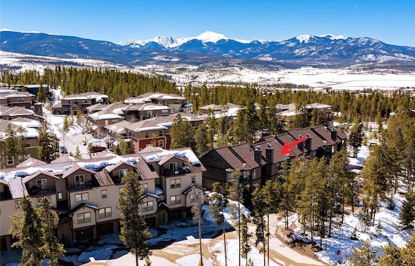 Área residencial com várias casas, algumas com neve nos telhados, cercada por árvores e montanhas ao fundo.