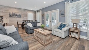 Un salon moderne avec un canapé gris, une table basse et une table d’appoint avec une lampe. On y trouve également un coin repas avec une table et des chaises, ainsi qu’un ventilateur de plafond.